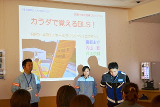 「カラダで覚えるBLS!」と表示されたスクリーンの前で、青いジャンパーを着た講師3人が受講者の前に立って挨拶を行っている写真