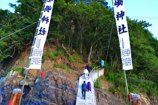 岩場の先に続く石段を参拝者が上っており、白い布に「荒嶋神社」と書かれた大きな旗が両脇に立っている神社参道の写真