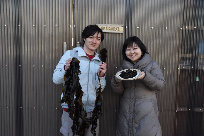 「金比羅丸」と書かれた小さな木の看板が設置された波板の壁の前で、長い生のわかめを両手で持った高橋さんと、ワカメが盛り付けられた皿を持ったリポーターの女性が並んで笑顔で立っている写真