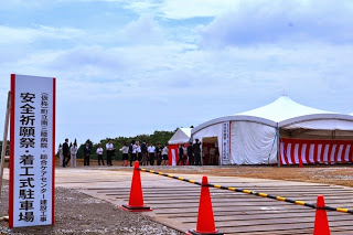 白いテントが張られた会場入口に「安全祈願祭・着工式駐車場」と書かれた看板が設置され、スーツ姿の人々が集まっている様子の写真