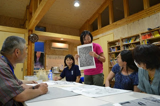 木造の室内で女性が切り絵の下絵を皆に見せながら説明しており、周囲の参加者が注目している写真