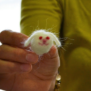 黄緑色の服を着た人が、作成した白い羊毛フェルト製のうさぎの顔を指でつまんでカメラに向かって見せている様子をアップで撮影した写真