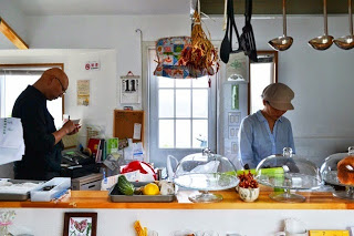 厨房スペースで女性が調理を行い、男性がレジ付近でメモを取っている、小さなカフェの営業準備の様子を写した写真