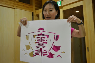 女性が木造の室内で「宝」と書かれた帆船のデザインの切り絵を両手で持ち、笑顔で完成作品を披露している写真