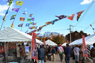 青空の下、色とりどりの旗が風になびく中、たくさんの人々がテントに囲まれた屋外イベントに参加している様子の写真