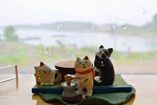 赤いマフラーをした三毛猫が餅をついている様子を、畳風のマットの上に置かれたこたつを囲む3匹の猫が見ている置物が並んでおり、窓際に置かれた背景にぼんやりと海が見えるユニークなインテリアの写真