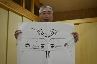 白髪まじりの男性が桜の花とお膳の図柄が切り抜かれた切り絵作品を胸の高さで広げて見せている写真