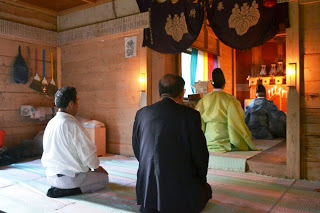 拝殿の中で祭祀服を着た2人の神職と2人の参拝者が、祭壇を向いて座り祈りを捧げている室内の写真