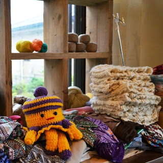 毛糸玉や毛織物が並べられている木製の棚の前に、手編みのぬいぐるみや布製品が積まれている店内の一角の写真