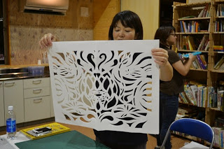 黒い服を着た女性が、大きくて複雑な模様の切り絵の作品を両手で持って皆に披露している写真