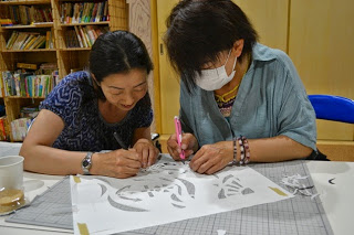 二人の女性が一枚の切り絵を一緒に作業しており、互いに協力しながら仕上げをしている様子の写真