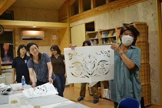 完成した切り絵を持った水色の服を着た女性が笑顔でポーズをとり、後ろに他の参加者も写っている写真