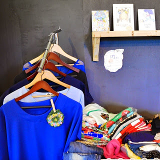 黒い壁に沿ってブルー系のシャツがハンガーにかけられ、下にはカラフルな服が山積みになっている写真