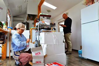 室内で女性が届いた段ボールを開けており、奥で男性がメモを取っている、小さな店舗内のキッチンの様子の写真