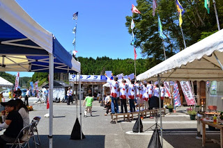 青空の下の地域イベント会場で、台の上に乗った白い法被姿の大勢の人が並んで両手を上げている写真