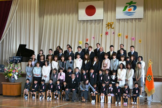 体育館の舞台前で、正装をした小学校の新入生とその保護者、教職員の方々が数列に分かれて並び、国旗と校章の垂れ幕の下で記念撮影を行っている入学式の写真