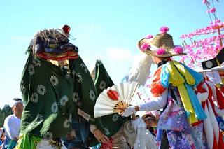 緑色の獅子舞が迫力ある表情で踊る中、色とりどりの衣装を着た踊り手が扇子を持って対峙している写真