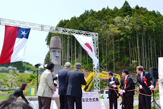 チリ国旗と日本国旗が上に掲げられた屋外の式典会場で、複数のスーツ姿の人々がモアイ像の前に立ち並び、赤いロープを引いてモアイ像のお披露目をした瞬間の写真