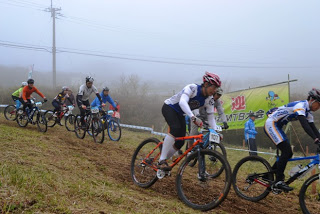 霧が立ち込める中、草地の坂道をヘルメットをかぶった複数の選手たちがマウンテンバイクで下っている「MTB大会」の様子の写真