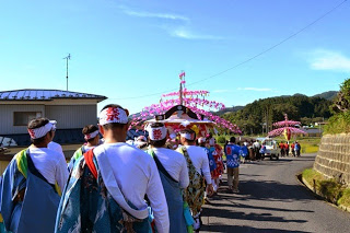 田畑が続く田舎道を、山車を囲むようにして祭り装束の人々が列を作り、ゆっくりと練り歩いている写真