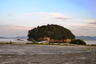 夕暮れ時の海岸から見た、木々が密集した小島が遠くに浮かび、手前には砂利と草地が広がる風景の写真
