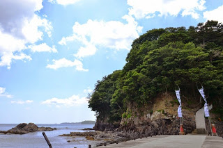 青空のもと海岸に面した小島の岩肌と緑が見え、手前に石造りの道が続いている海岸線の写真
