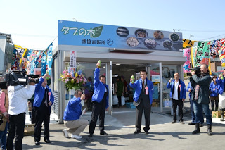 テレビカメラによる撮影が行われる中、「タブの木」と書かれた看板の店舗の前で青いジャケットを着た数人がこぶしを握った手を挙げている、漁協直販所のオープンの様子の写真