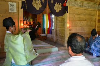 神社の拝殿内部で祭祀服の神職が両手で榊を持ち、隣に正装した参拝者たちが座っている祭祀の様子の写真
