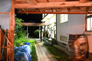 夜の校舎の外で、明かりのついた窓から中の様子をうかがう数人の人々がいる様子の写真