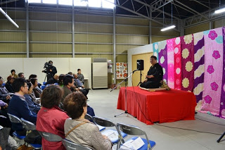 色とりどりの布が飾られた背景の前で、赤いクロスがかけられた台に座った落語家の男性がマイクを使って落語を披露している様子の写真