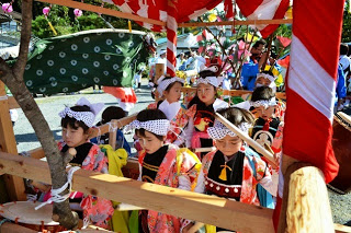 子どもたちが揃いの鉢巻きを巻き、華やかな衣装を着て山車の中で真剣な表情で太鼓を叩いている写真