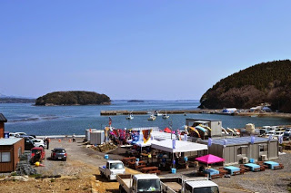雲一つない晴天の空の下、仮設の建物やテントが並ぶ漁港の前に海が広がり、小さな島々と停泊する船が見える写真