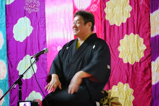 ピンクや紫色などのカラフルな布に大きな黄色と白の花柄が描かれた背景の舞台で、黒い着物を着た落語家の男性がマイクの前に座って落語を披露している様子の写真