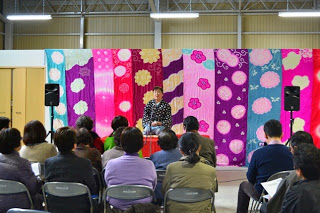 色とりどりの模様が描かれた布で飾られた背景の舞台で、和装姿の仲松先生が話しており、椅子に座った観客が見ている様子を後方から撮影した写真