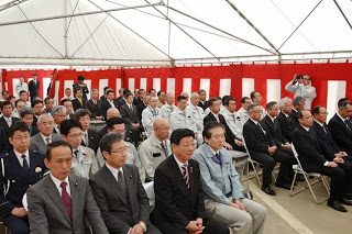 白い屋根と紅白幕の壁を持つ大きなテントの中で、制服やフォーマルな服装をした関係者が整然と座っており、右側にはイベントの様子を撮影している人物がいる終了祭の様子の写真