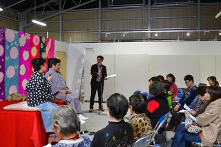 色鮮やかな布が背景に飾られ、赤いクロスがかけられた小さなステージに2人が座っており、右側のマイクスタンドの前に立つ男性とステージに座っている女性が話をしている様子を、集まった数人の観客が椅子に座って聞いている写真