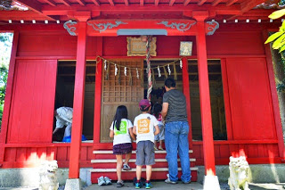 赤い神社の建物の前で、子供3人と大人1人が鈴を鳴らしお参りをしている後ろ姿が見える写真