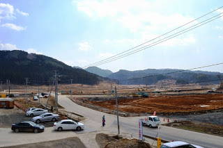 背景には山々と雲のある青空が広がり、道路脇に車や白いトラックが停まっており、右側の建設予定地の地面が平らにならされて工事の準備が進められている建設現場の写真