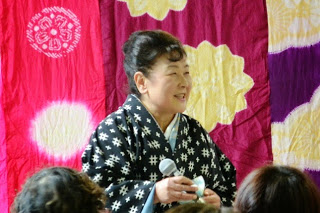 赤・黄・紫の模様がある色鮮やかな背景の前で、和装姿の笑顔の仲松先生が両手でマイクを持ち、複数の人々がその人物の方を向いている様子の写真