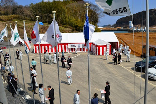 側面に紅白幕が張られたテントの前に多くの人々が集まり、掲揚台に複数の旗が立てられている、終了祭の様子の写真