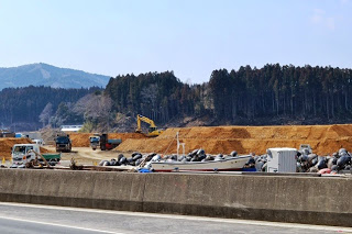 木々と丘に囲まれ、トラックやダンプカーが停まっている建設予定地に土の山があり、ショベルカーによる重機で作業が進められている建設現場の写真