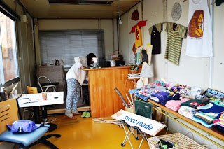 木製のカウンターの後ろに一人の人物が立って何か作業をしている様子が見られ、壁には衣類が掛けられ、テーブルの上には折りたたまれた衣類が並べられていて、店内には看板や雑貨などが散りばめられている写真