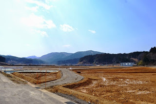 青空と雲が広がり、背景に遠くの森林に覆われた丘や山のふもとに建物が見え、乾いた土が広がる開けた野原に曲がりくねった未舗装の道が通っている建設予定地の写真