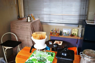 手前にはガラス容器の上にフィルター付きのコーヒードリッパーが置かれ、その隣には黒い手動式コーヒーミルと瓶が並び、奥には布をかけたテーブルの上に電気ケトルや小物が置かれていて、壁際の棚には本や装飾品が並び、窓にはブラインドが閉じられている写真