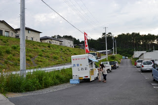 赤いのぼりが立っている移動販売車が坂のある住宅街の道路わきに停まっており、数人の客が並んでいる様子を写した写真