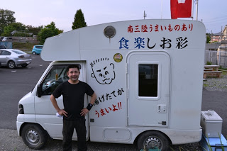 車体に「食楽しお彩」「揚げたてがうまいよ!!」と手書き風の文字と顔のイラストが描かれている白い軽バンの移動販売車の横に笑顔の店主後藤さんが立っている写真