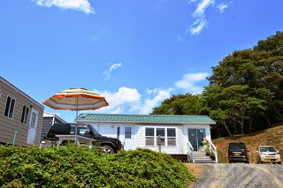 青空の下、丘の上に建つ緑色の屋根で白いプレハブ風の建物の前に車が数台停まっており、カラフルなパラソルが立てられた開放的なカフェの外観の写真