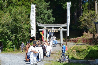 鳥居をくぐって白装束の神職や氏子たちが旗を持って境内から道へと進んでいく行列の写真