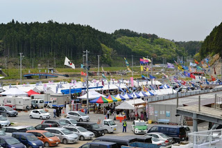 緑の森と山を背景に、たくさんの白いテントや色とりどりの屋根付きテントが立ち並び、色鮮やかなのぼり旗が風になびいているイベント会場全体の写真