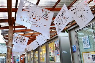 木の柱と透明な波板の屋根がついた通路に、細かく切り抜かれた切り絵が吊るされている商店街の通りの写真